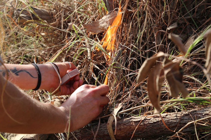 A close up of a woman's hands holding a box of matches as a small fire starts to burn in the bush.