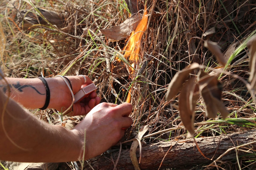 A close up of a woman's hands holding a box of matches as a small fire starts to burn in the bush