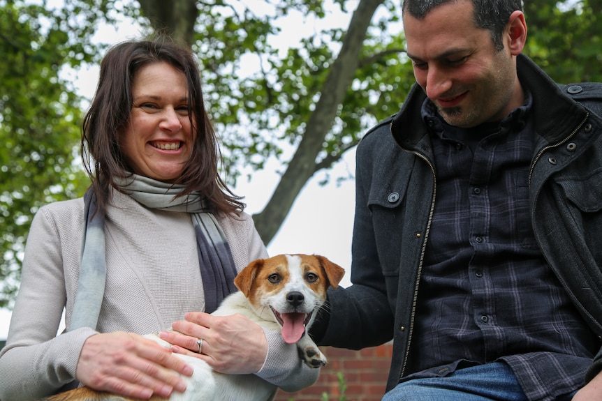 A man and a woman hold a small brown and white Jack Russell on her lap.