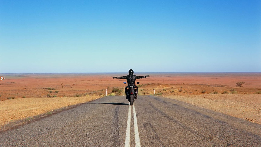 A motorbike rider with arms outstretched riding along the double lines on a desert road.