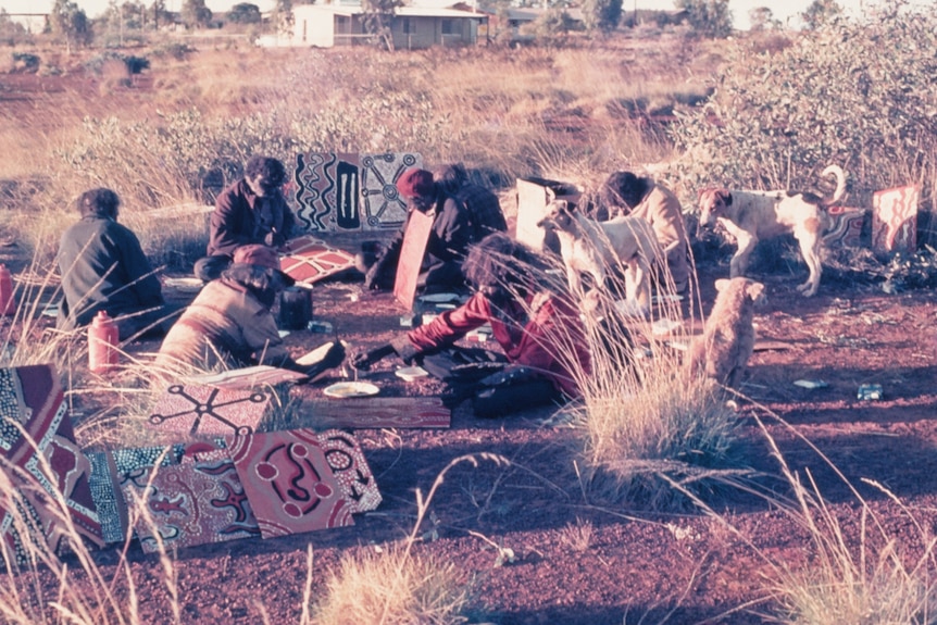 A group of First Nations men sit amongst spinifex clumps and dogs painting