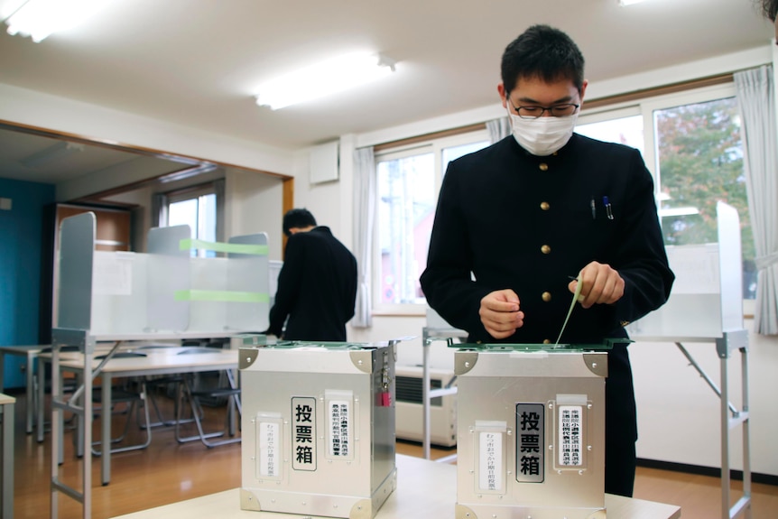 A man in a black suit puts paper in a ballot box.
