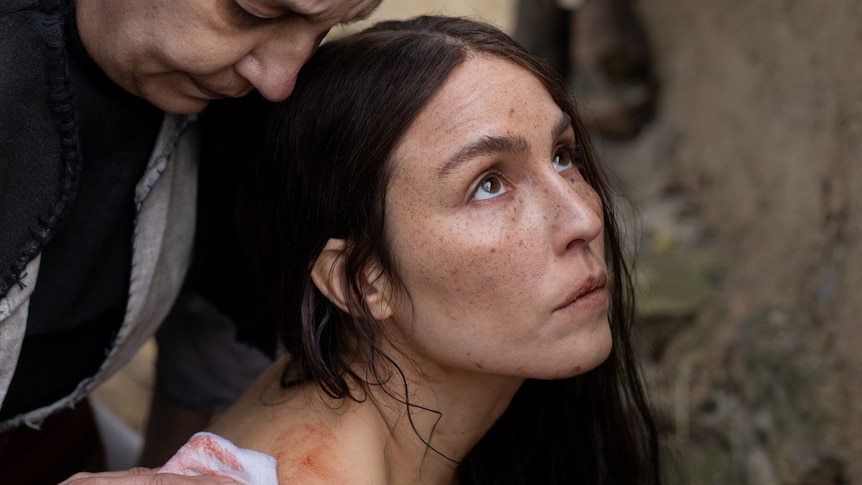 A woman with long dark hair, brown eyes and freckles looks up while an older woman looks over her shoulder