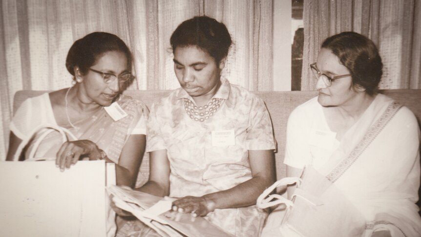 Three women sit on a couch, two are wearing saris.