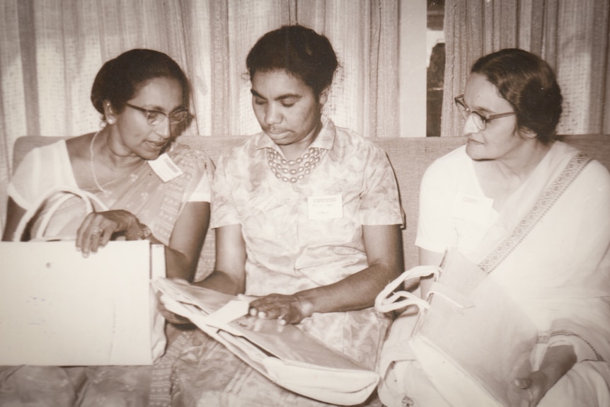 Three women sit on a couch, two are wearing saris.