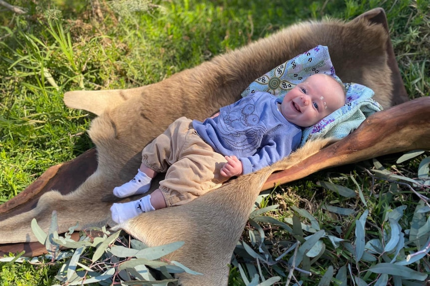 Baby smiles while lying on a kangaroo skin. He is wearing a blue jumper and light brown pants.