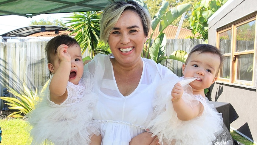 Woman with short blonde hair in a flowy white dress holding two small children in both arms wearing white leotards, trees behind