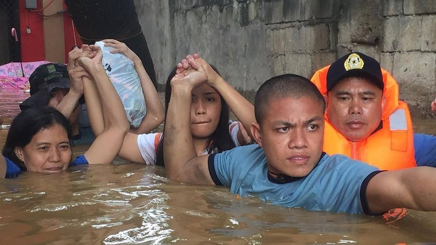 Three police officers help hold the hands and belongings of two women in brown floodwaters up to their necks