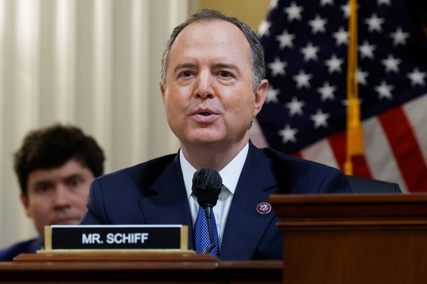 representative adam schiff speaks into a microphone while sitting at a desk behind a nameplate