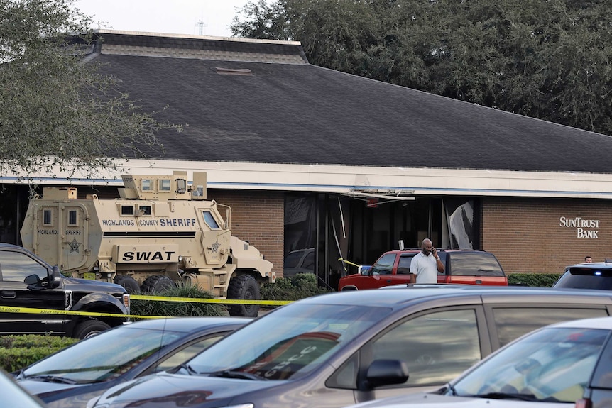 A beige SWAT vehicle is seen in front of a brown brick bank with cars in the parking lot seen in front.