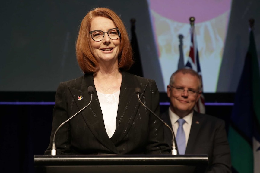 Julia Gillard stands at a podium smiling out at the audience, while Scott Morrison looks on from behind, also smiling.