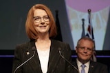 Julia Gillard stands at a podium smiling out at the audience, while Scott Morrison looks on from behind, also smiling.