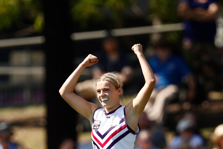 An AFLW footballer grins through her mouthguard and raises her arms in triumph after kicking a goal.