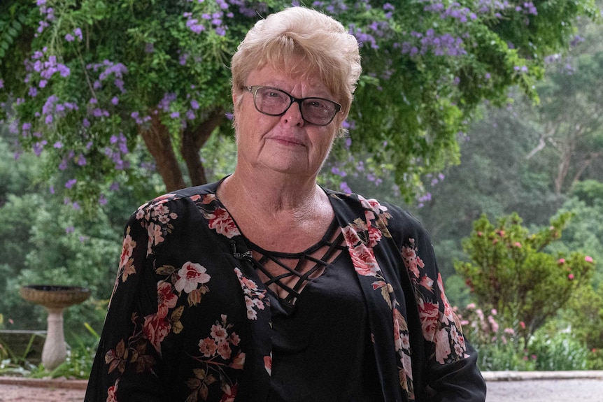 Heather Slade wears a black and floral top, wearing glasses. Behind her is a lovely green garden with purple flowers.