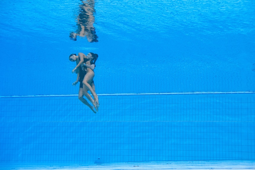 A woman, carrying an unconscious woman, swims from the bottom of a pool to the surface.
