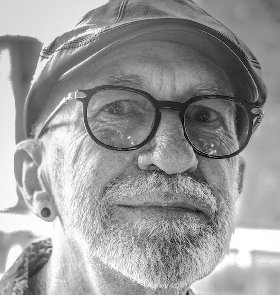 A black and white photo of a man in his 70s with glasses and a cap.