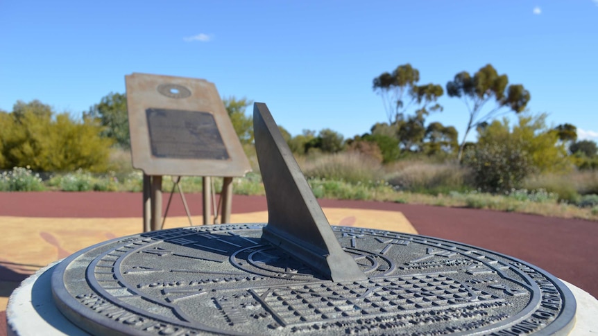 A metal sundial in the foreground, behind it, a steel sign in a courtyard surrounded by arid plants under a bright blue sky.