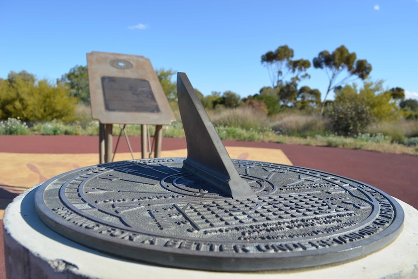 A metal sundial in the foreground, behind it, a steel sign in a courtyard surrounded by arid plants under a bright blue sky.