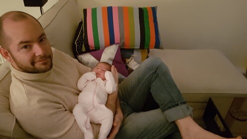 Matthew Patrick sits on a couch with a newborn baby.