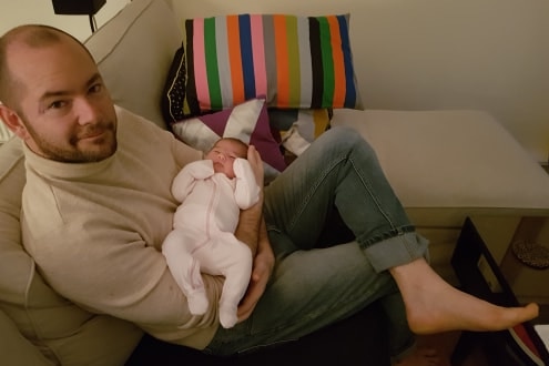 Matthew Patrick sits on a couch with a newborn baby.