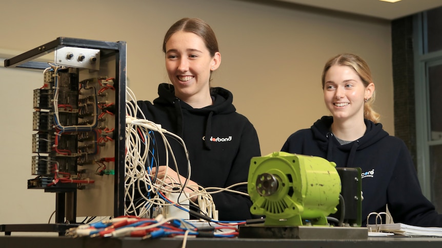Lucy Cliff and Lauren Blasi smile while plugging cords into a panel. They both wear dark hoodie jumpers.