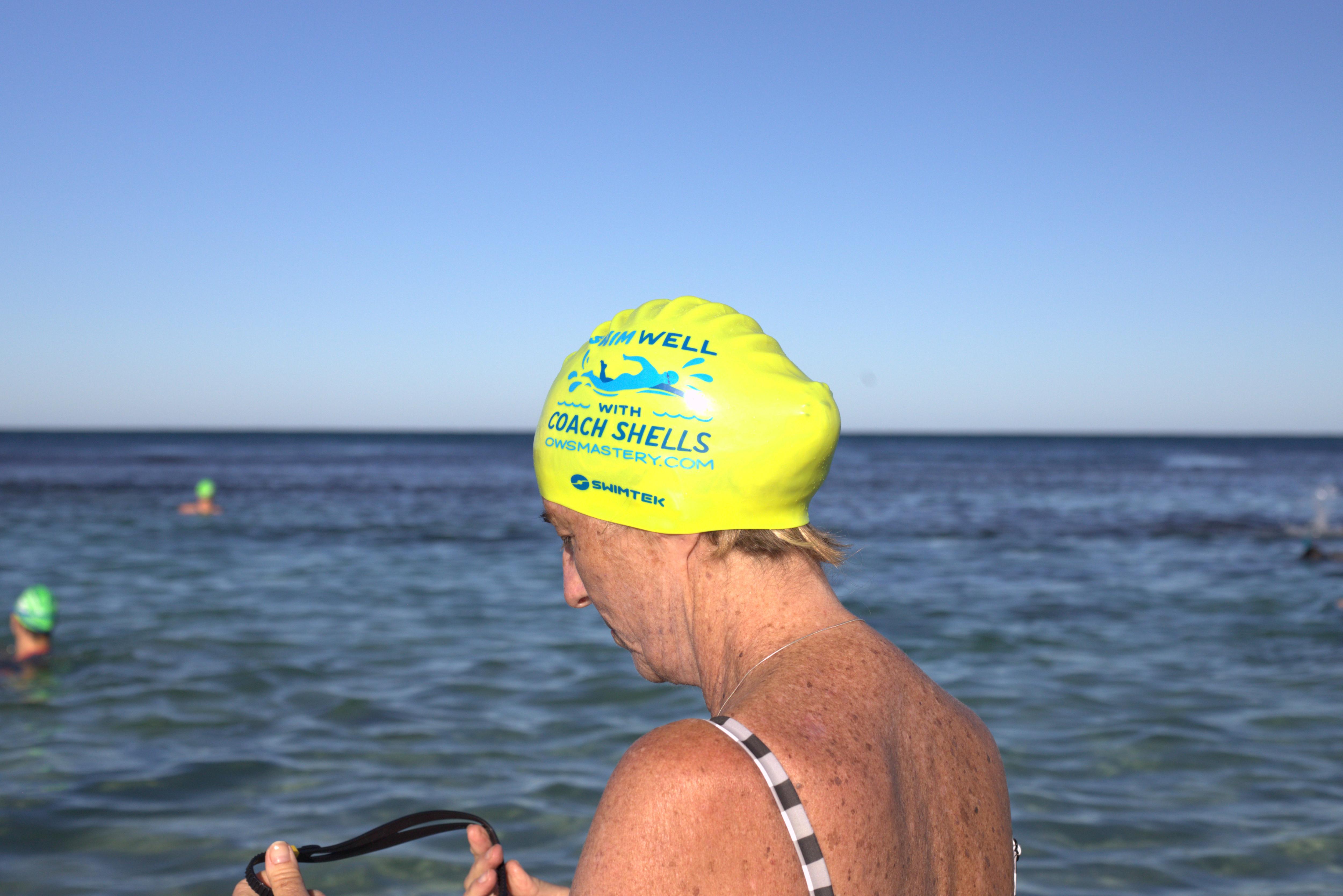 A swimmer gets ready for the water, she wears a yellow "swim well with coach shells" swim cap.