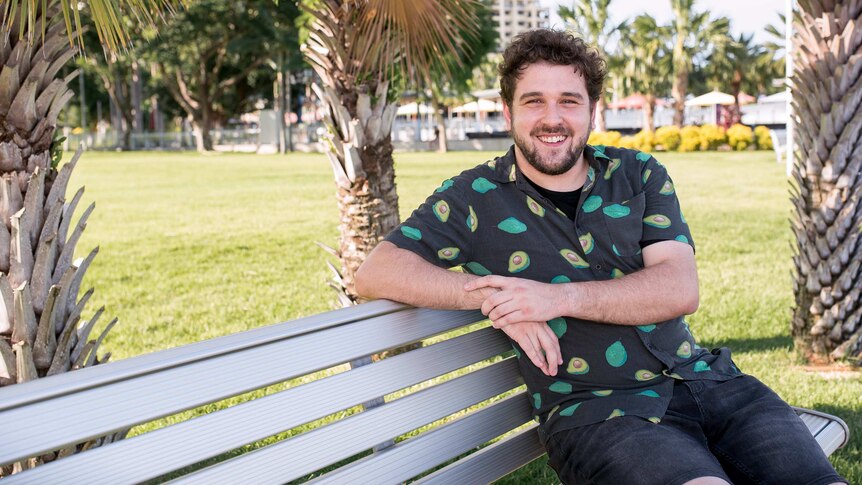 A man with dark curly hair and a beard, wearing a shirt with an avocado print,  sitting on a park bench.