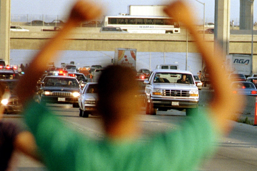 A blurry photo of a man in the forefront with a highway car chase in the background, a white SUV and police cars behind it.