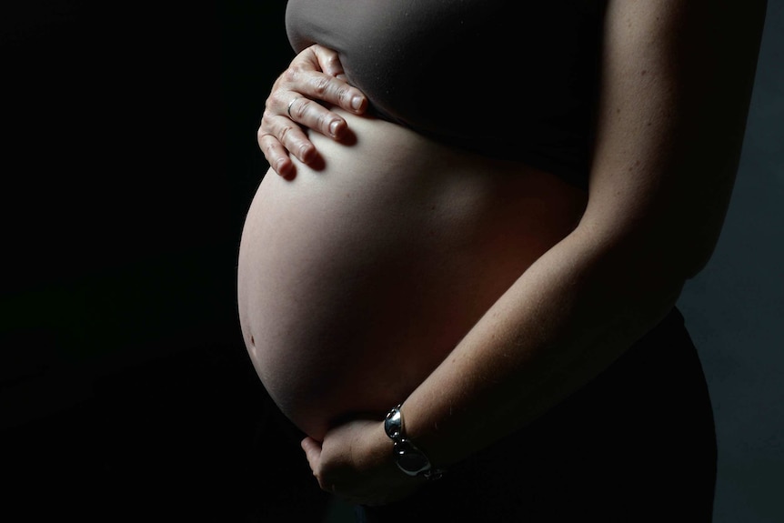 A pregnant woman's torso stands out against a dark background. She has one hand perched on the top of her bare belly.