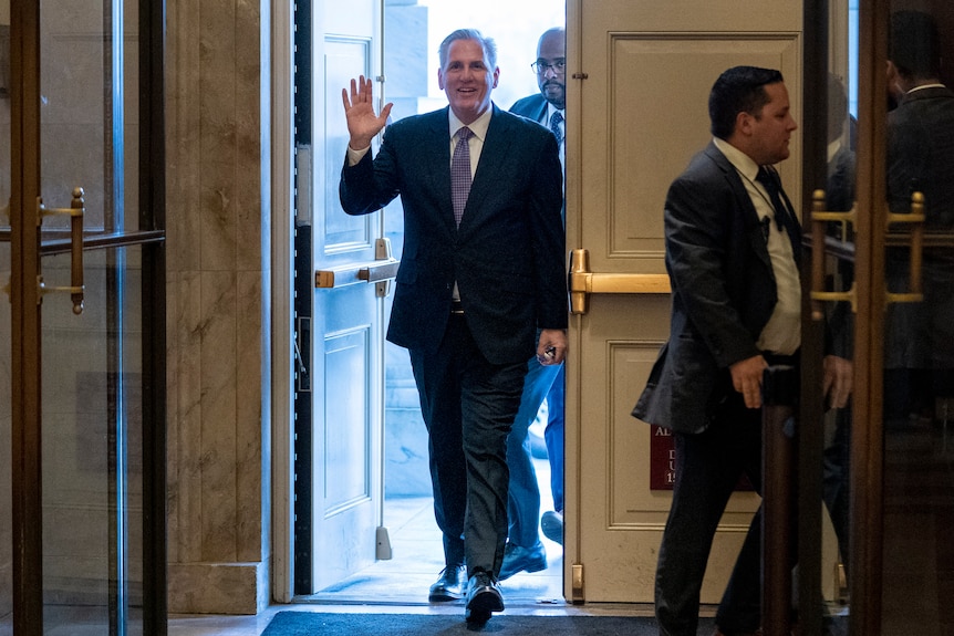 A man in a suit walks through the door of Congress waving.  