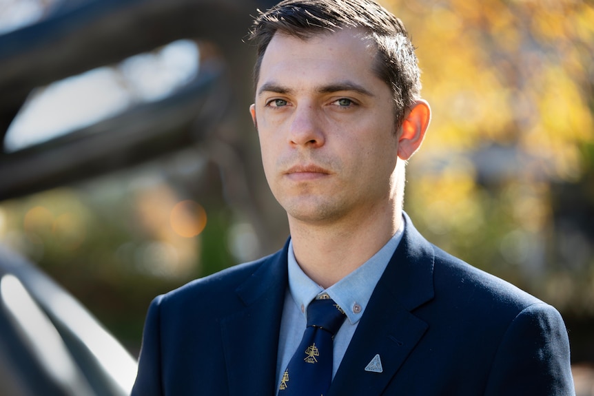 Man in a blue shirt and navy jacket wearing a tie, looking serious.