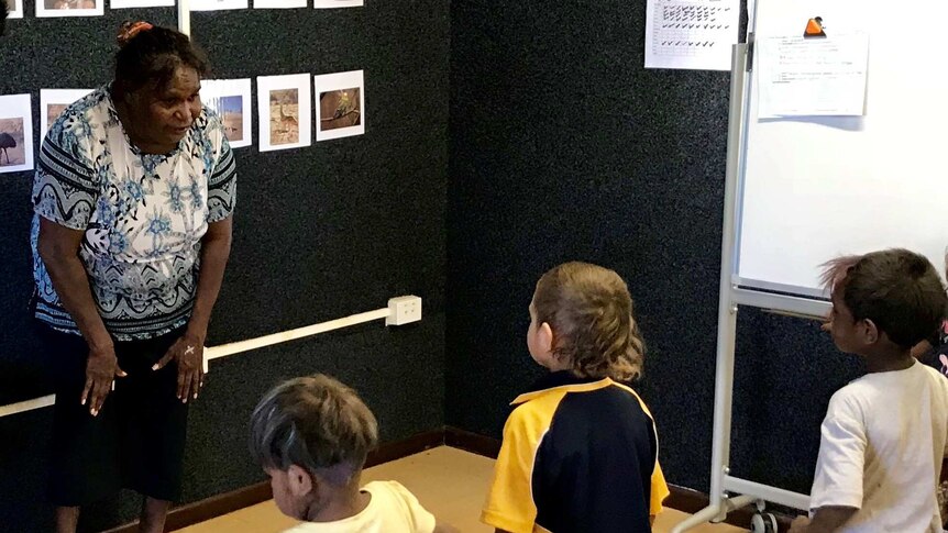 Three small children listen to an Indigenous Australian woman in a classroom setting