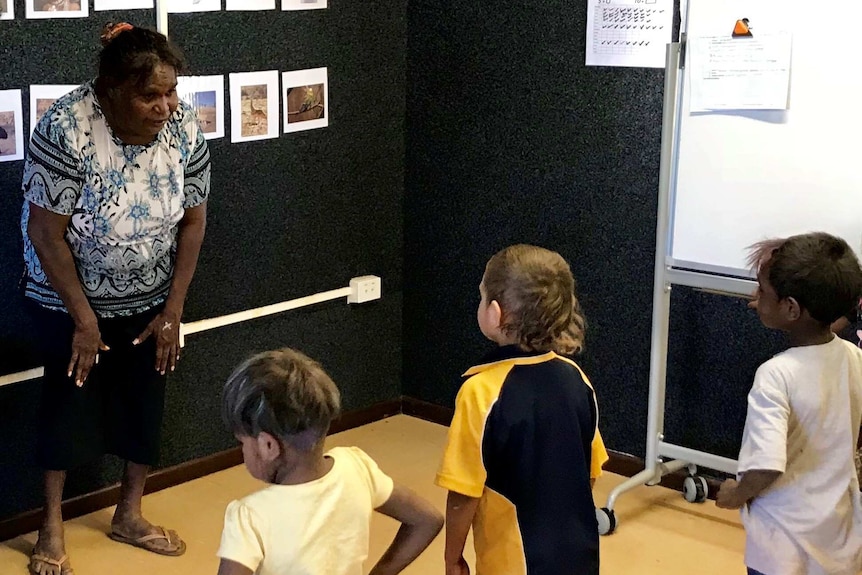 Three small children listen to an Indigenous Australian woman in a classroom setting