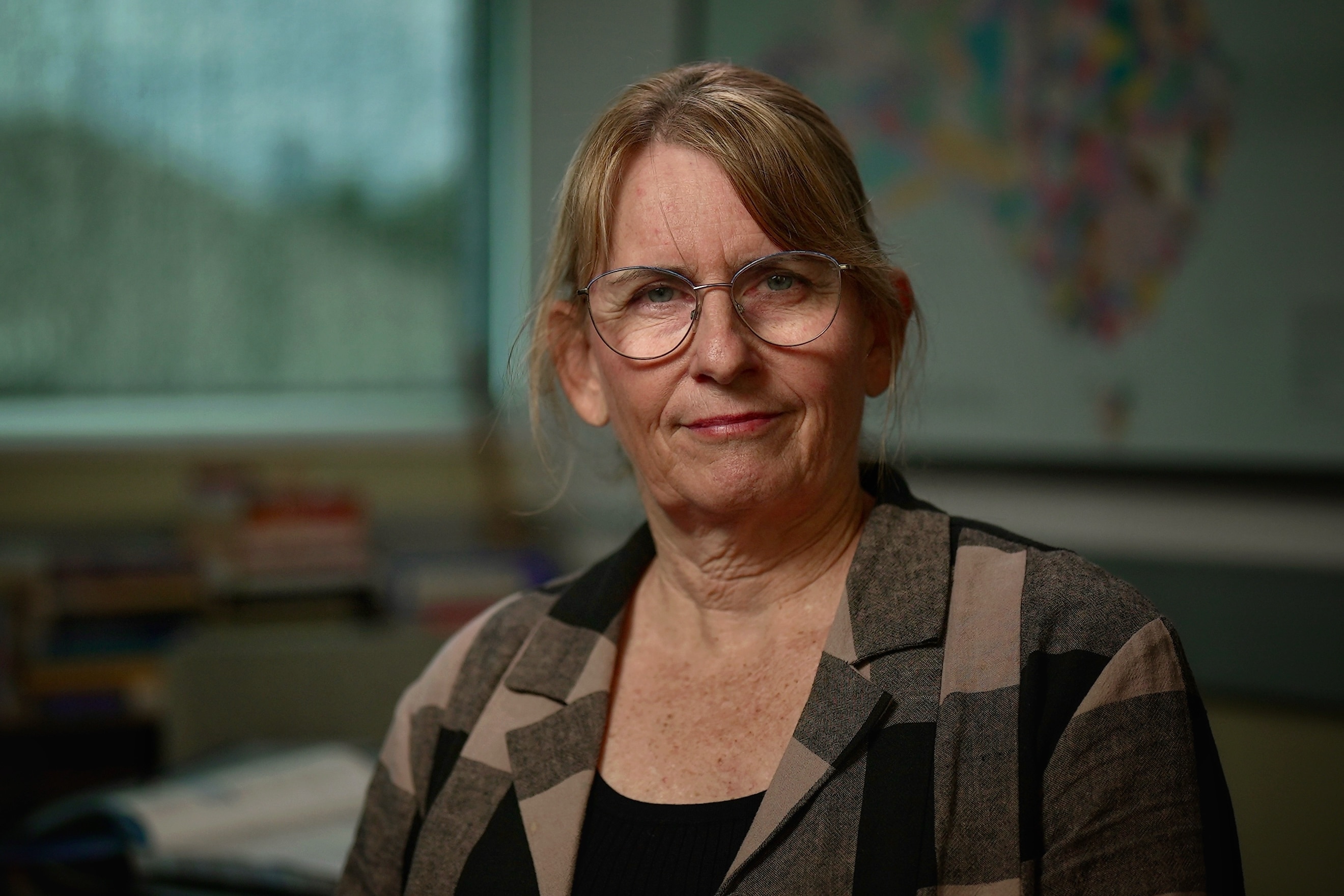 A white woman with blonde-brown hair tied back, wearing glasses and checkered jacket sitting in office looking at camera.