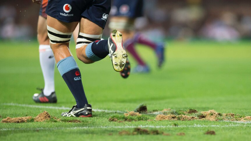 A rugby player runs over a grass surface with significant divots