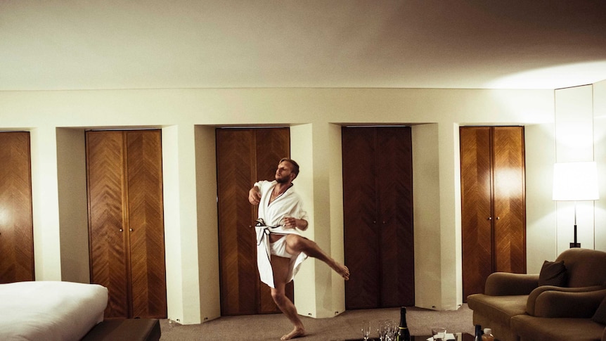 A man in a dressing gown and white underwear dances in a modern hotel room