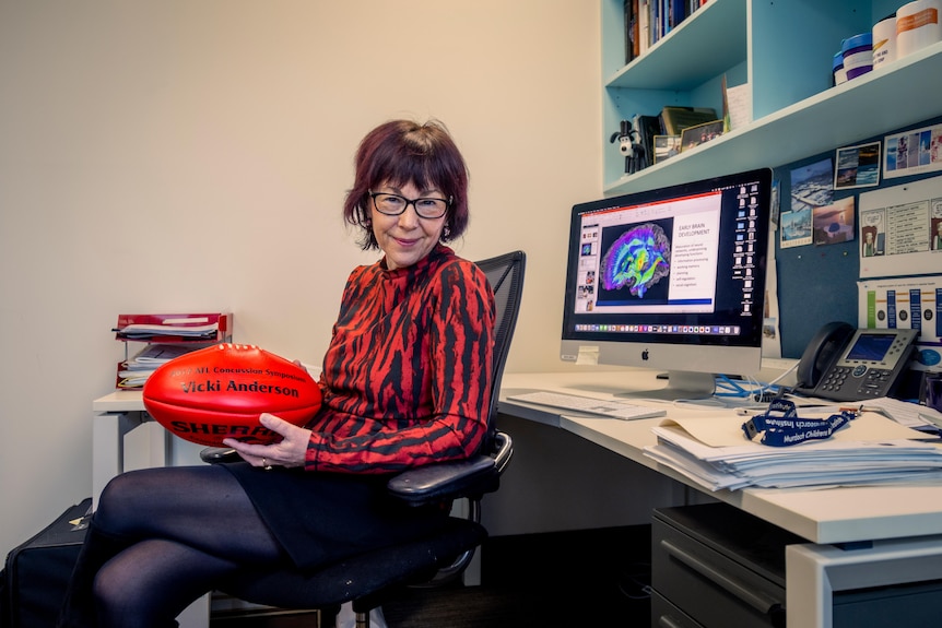 A woman wearing a black and red striped shirt sits in an office, holding a red football.