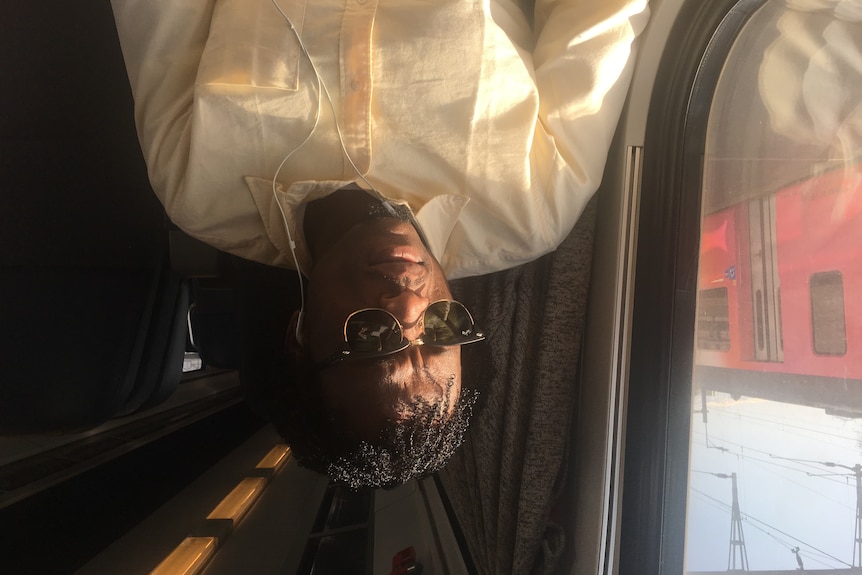 Man wearing sunglasses and light shirt sits next to a window on a train.