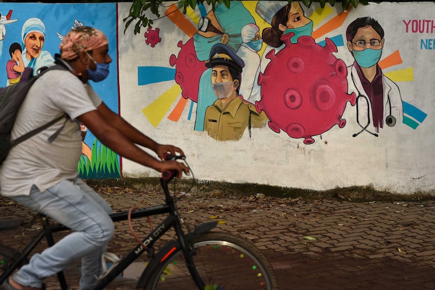 A man wearing a mask pedals his bicycle past a wall with graffiti
