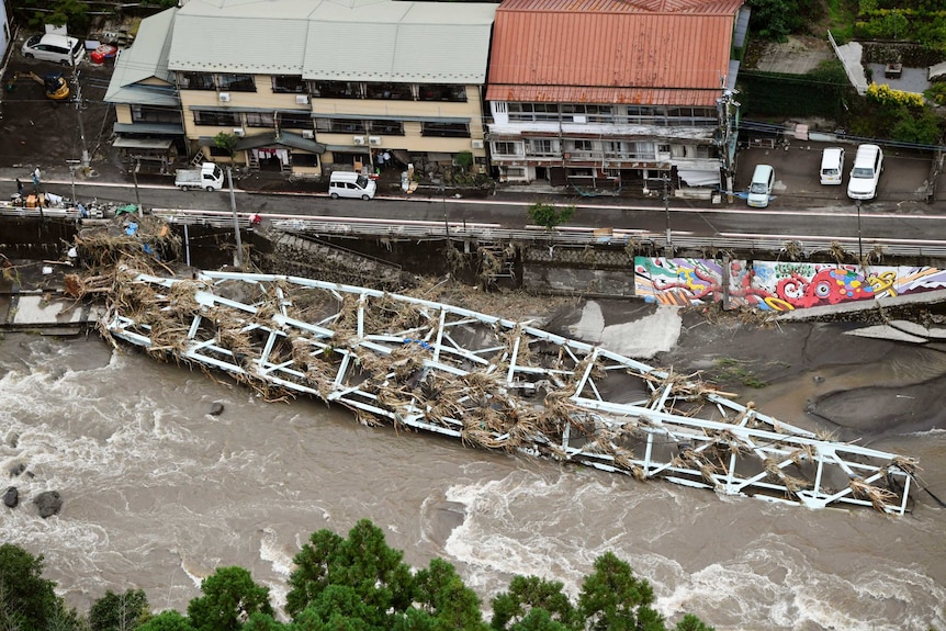 An aerial photo shows a small bridge strewn with debris that has collapsed into a flooded river near residential buildings.