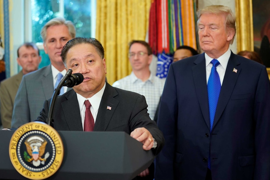 Hock E Tan stands at a lectern bearing the US presidential seal while US President Donald Trump looks on.