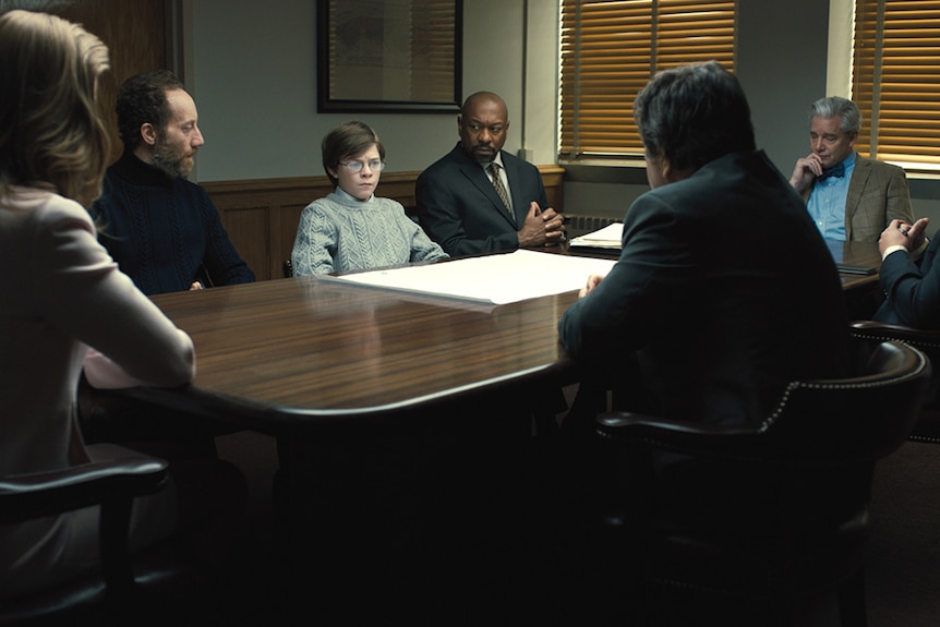 A young bespectacled boy sits at meeting table surrounded by adults in a room with closed curtains.