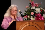 A woman stands at a lectern surrounded by flowers and next to a photo of her daughter.