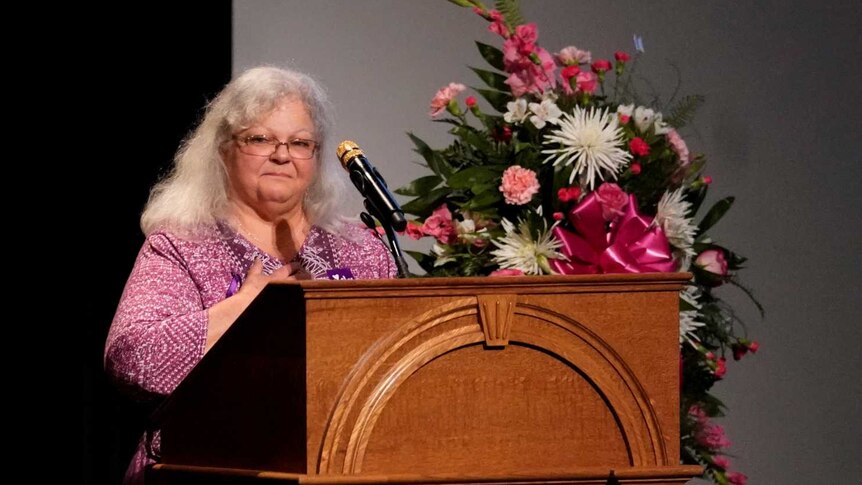 A woman stands at a lectern surrounded by flowers and next to a photo of her daughter.