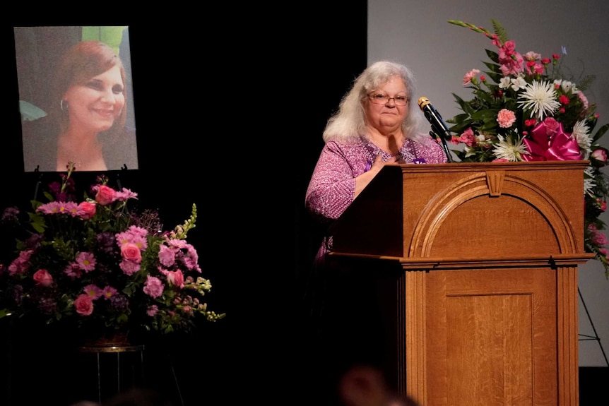 A woman stands at a lectern surrounded by flowers and next to a photo of her daughter.