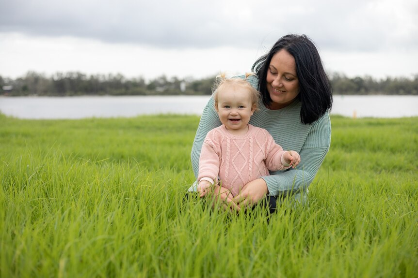 Tanisha Cowin hugs and smiles at a little girl wearing a pink jumper, in a green grassy field.