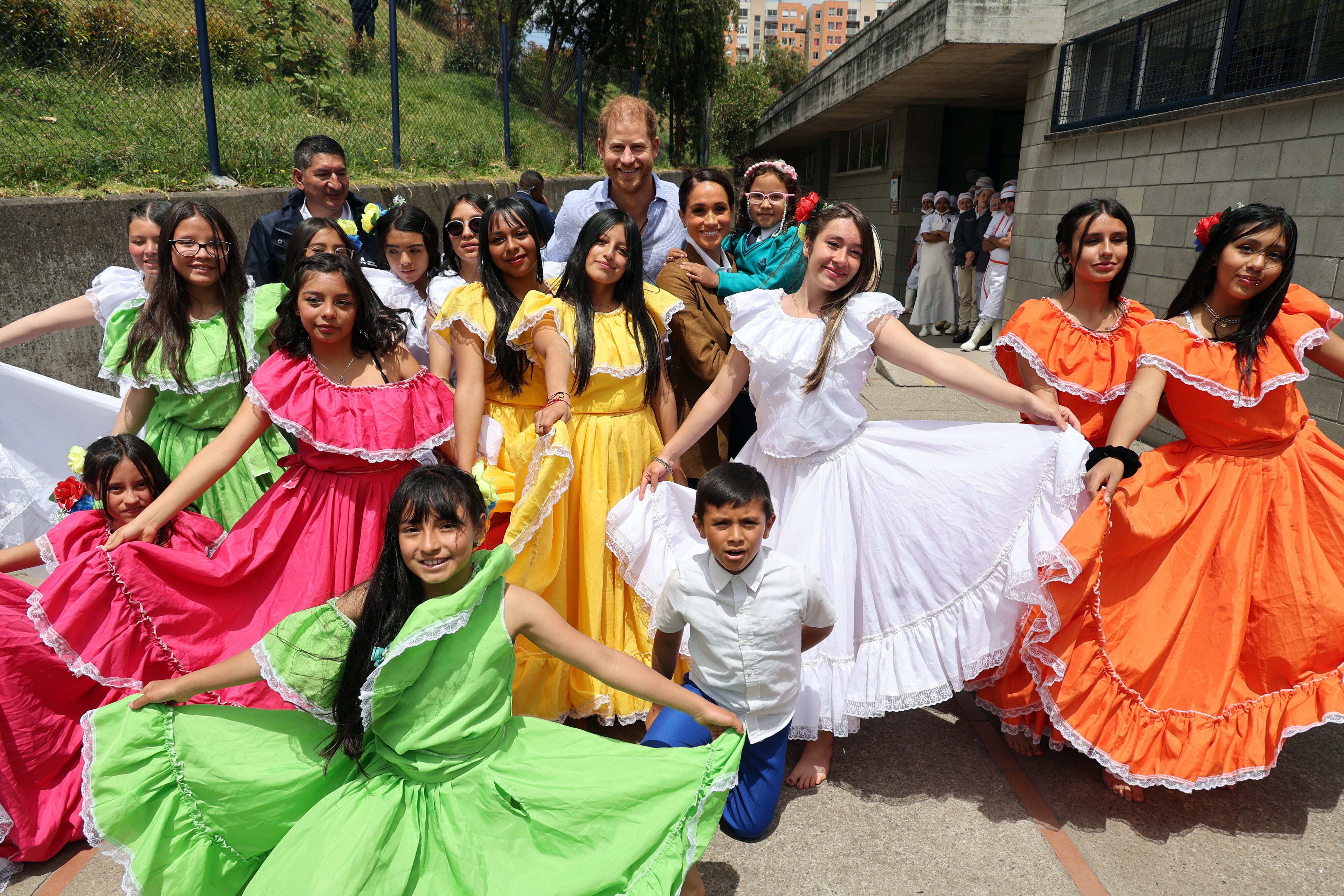 Harry and Meghan surrounded by a dozen students of different ages, dancing in bright traditional costumes.