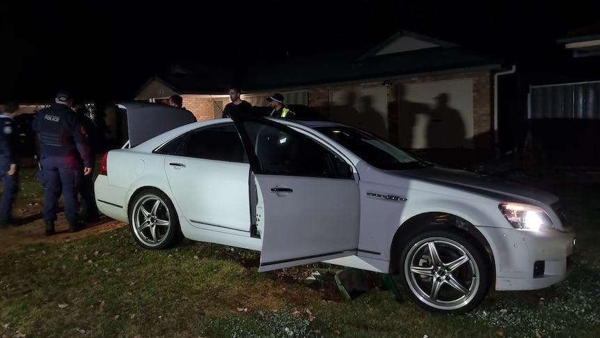 A large group of police officers gathered around a white Holden sedan at night.