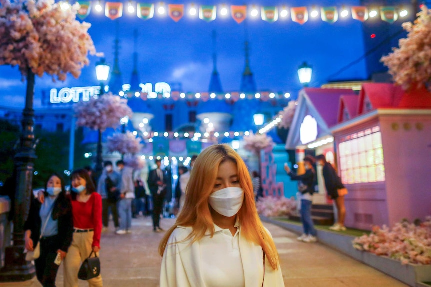A young woman in a face mask walks through a street strung with fairy lights in Seoul