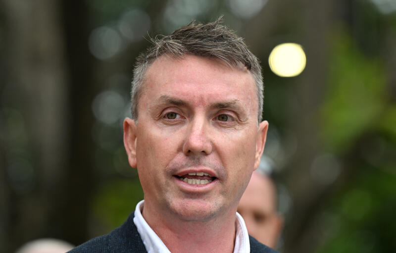 a close-up image of a white man speaking with green shrubbery in the background